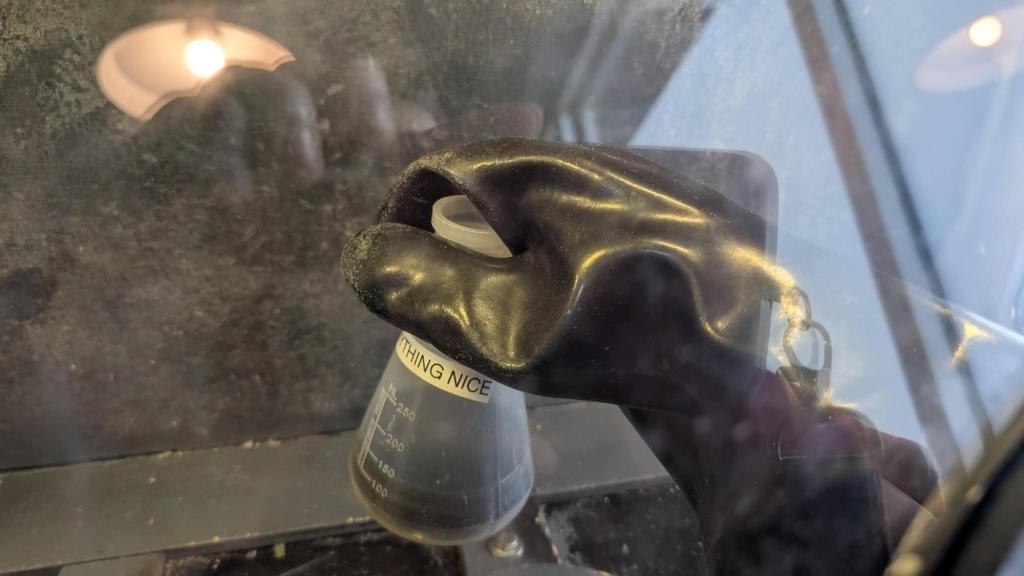 A black plastic gloved hand holding a clear beaker labeled "something nice." This is all instead of a plastic box.