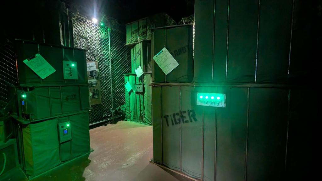 A fenced-in area with many stacks of large crates. Some have electronic panels, some have dangling clipboards with a form. There are spray-painted words on the side such as Fox or Tiger.