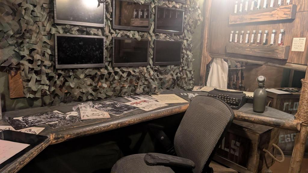 A desk attached to a wall, with some papers scattered across it and 6 monitors on the wall. Off to side is a typewriter, test tubes, crates and other paraphernalia.