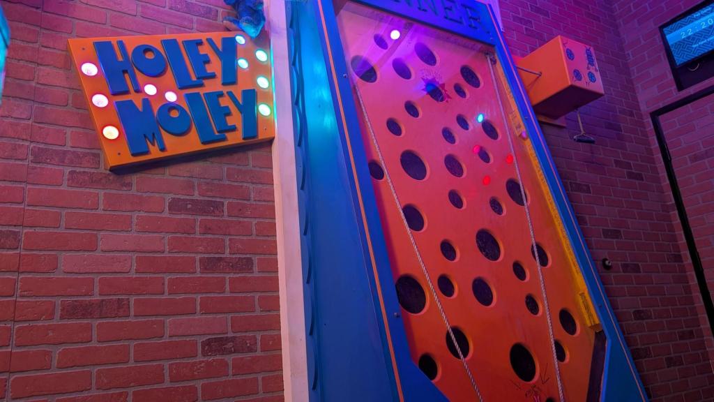 A blue and orange sign saying Holey Moley against a brick wall background. Next to the sign is a carnival games in the same colors where players lift game pieces with ropes and avoid dropping them in holes.