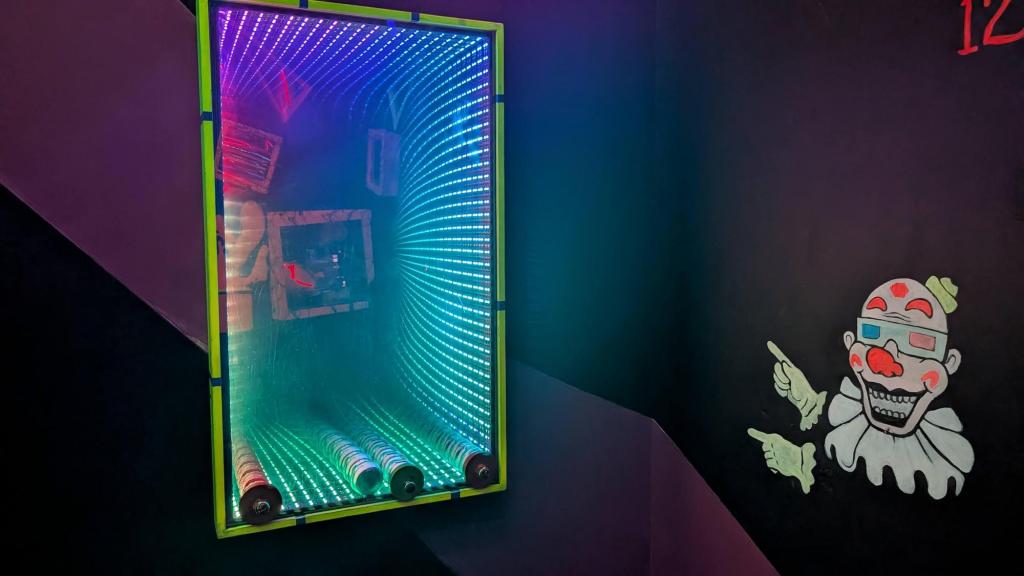 A view into a vortex of sorts with bright LEDs through a window set against a dark wall. On the wall is a painting of a clown pointing at the vortex window.