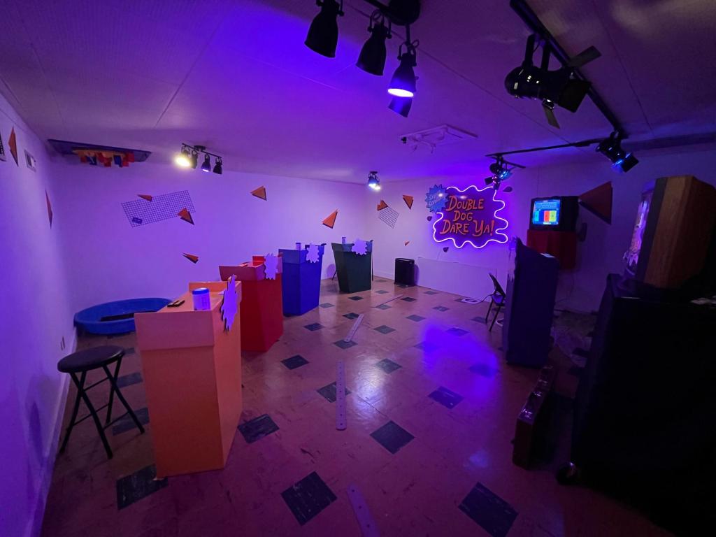 Wide-angle view of a room with orange and black linoleum floor and four colorful podiums arranged to face the emcee's podium. There's a neon Double Dog Dare Ya sign on the back wall.