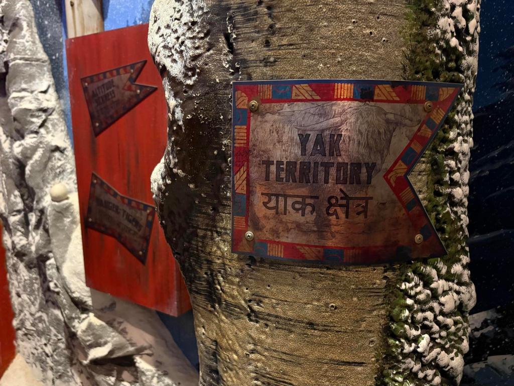 Closeup view of a snow-covered tree trunk with a colorful pennant-shaped sign. The sign says Yak Territory and a line of strange characters. Next to it sits a red wooden board with additional pennant-shaped signs but they are too far away to read clearly.