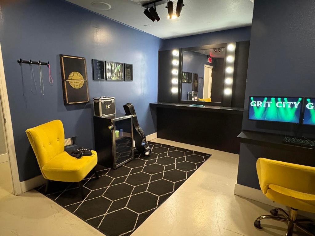 View of a room with blue walls, a large lighted mirror, and a mini fridge with a guitar case leaning on it. Two bright yellow chairs sit on a black, patterned carpet.