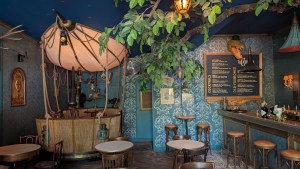 An ornate restaurant and bar area with patterned blue wallpaper, a hot air balloon or airship off in one corner, and a tree branch peeking into the frame.