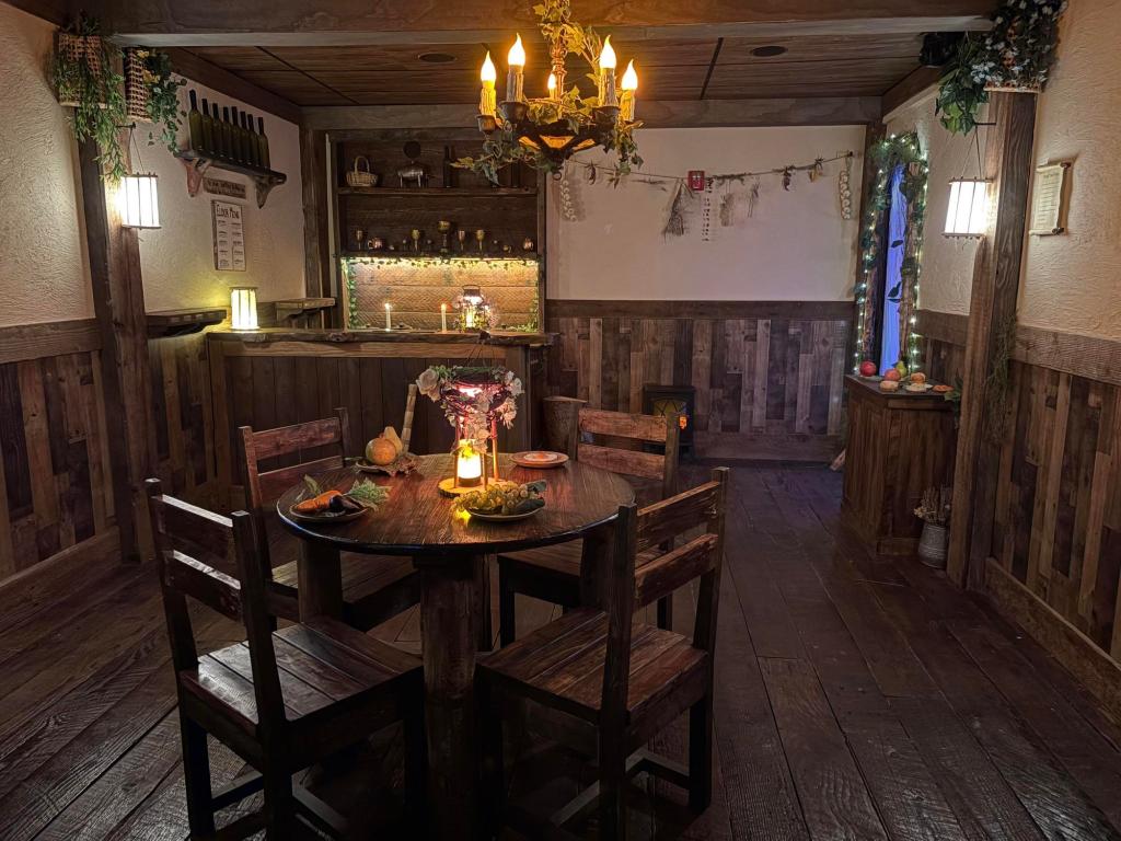 View of a cozy medieval tavern with wood floors, a round wooden table with four chairs, and a wooden bar at the back of the room. A chandelier and lanterns are lit up around the room, and the table is set with fruits on plates.