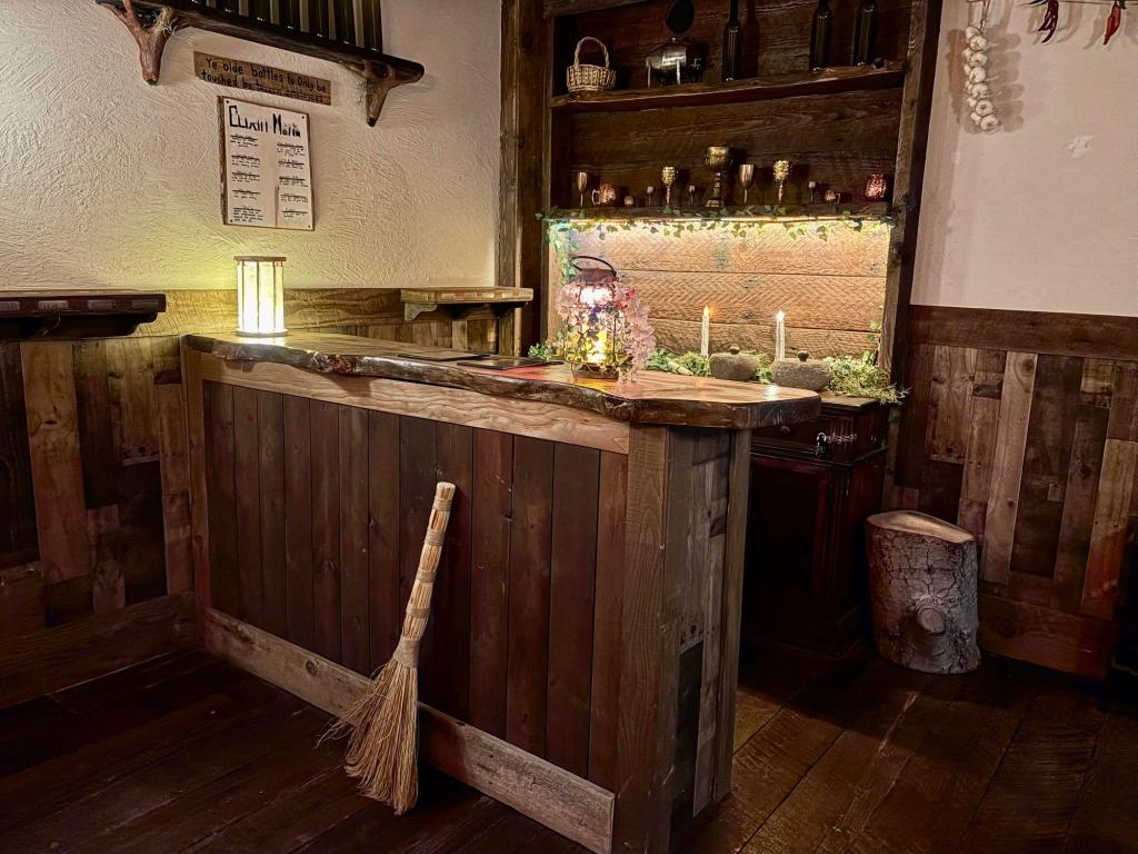 Closer view of a wooden bartender's bar, with goblets and bottles decorating the shelf behind it. Lanterns light up the space. A wooden shelf with a row of green wine bottles is on the left wall, while a straw broom sits propped against the front of the bar.