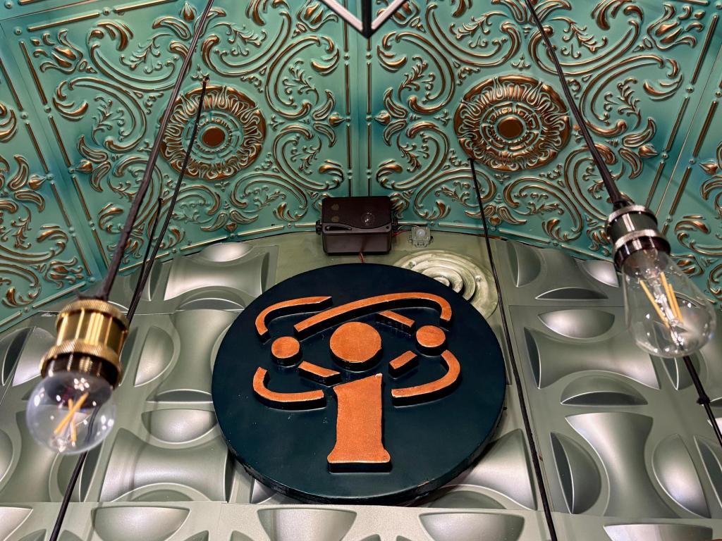 Image of an orange scientific logo on a circular black background. A lightbulb hangs down on either side. Intricate detailing at interest to the silver metallic wall and green and gold metallic ceiling near the logo.