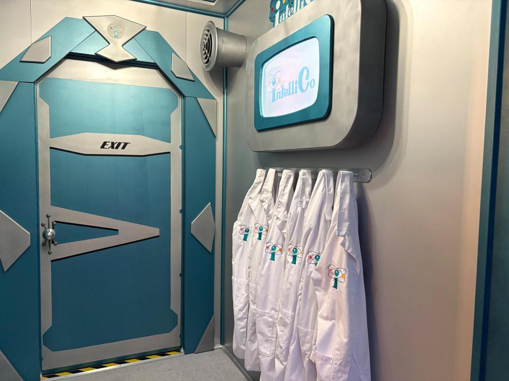 Small silver and blue futuristic room, with a retro TV monitor on the wall. Six white lab coats with the IntelliCo logo hang below the monitor. An Exit door is visible on one of the walls.