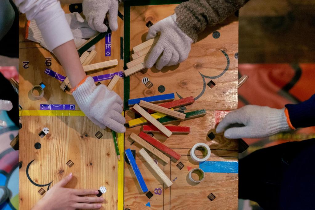 A square of wooden boards with various markings, and an array of white-gloved hands touching various colorful pieces.