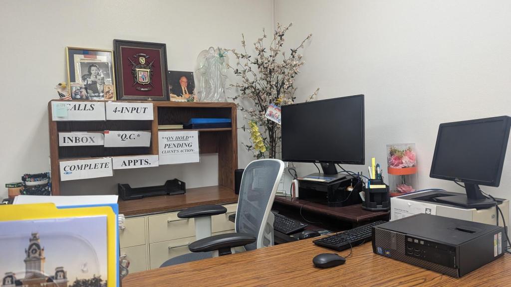 A desk area with computer equipment, with a shelf for client paperwork, which also holds framed photos and a family crest.