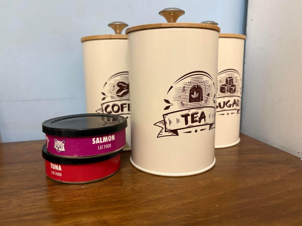 Three ivory canisters that say Tea, Coffee and Sugar sit on a wooden counter. Two cans of cat food, one salmon and one tuna, are next to the canisters.