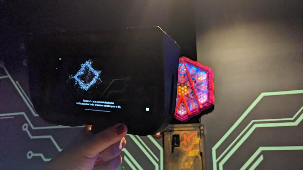 A hand holds a device with a screen showing that it is a particle detector. It is pointed at a colorful hexagonal collage mounted on the wall