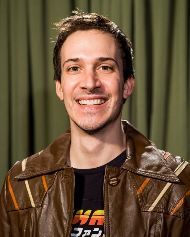 man with brown hair, slight stubble, wearing a brown leather jacket, smiling