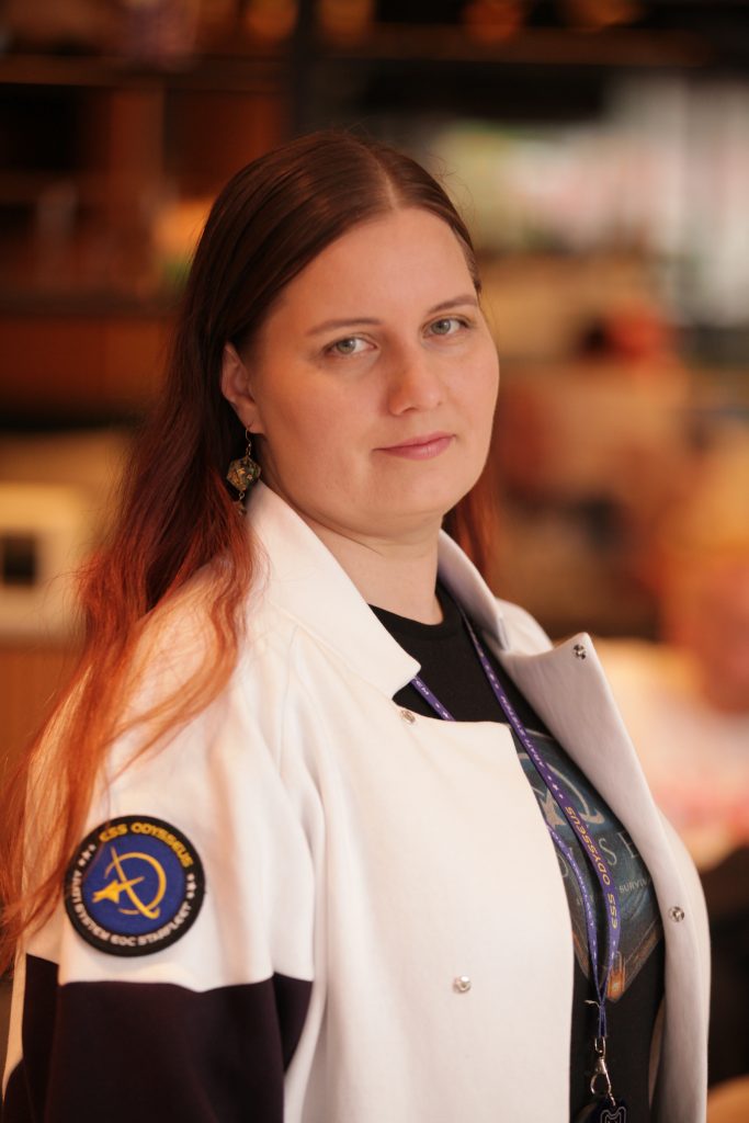 woman with long light brown hair parted in the middle, light grey eyes, wearing a white jacket with a odysseus patch on the arm