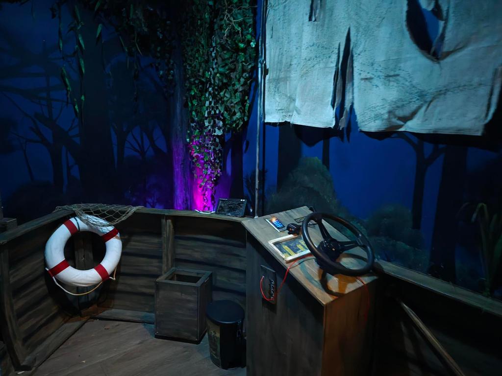 The backdrop is a mural of trees and bushes, with some vines and greenery hung from the ceiling. Standing on the deck of a boat next to steering wheel, navigation screens, and tattered sail.
