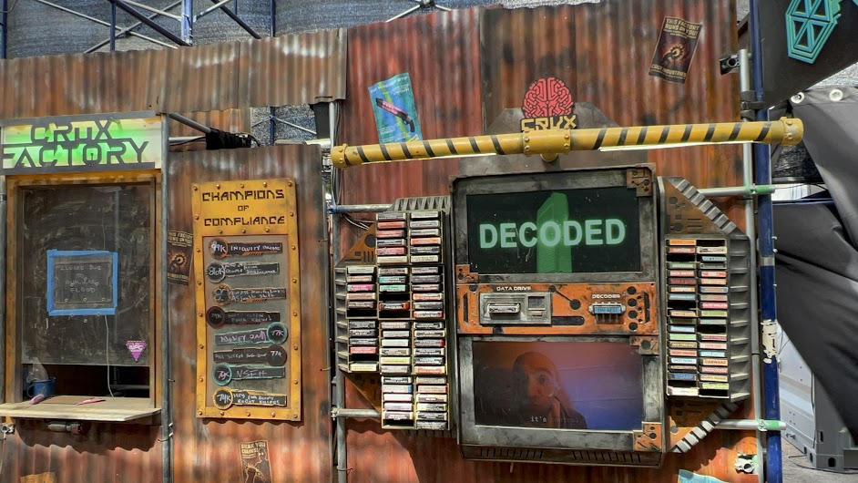 rusted looking corrugated metal walls with service window and sign that says "crux factory", decoder machine mounted on walls. board titled "champions of compliance"