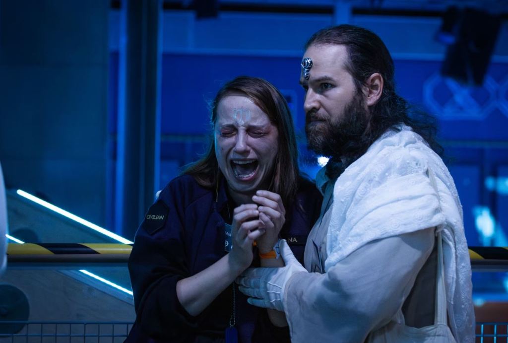 man in flowing white garments with a silver medallion on his forehead and long hair tied back, looking serious and holding a woman with long light brown hair who is crying in distress.