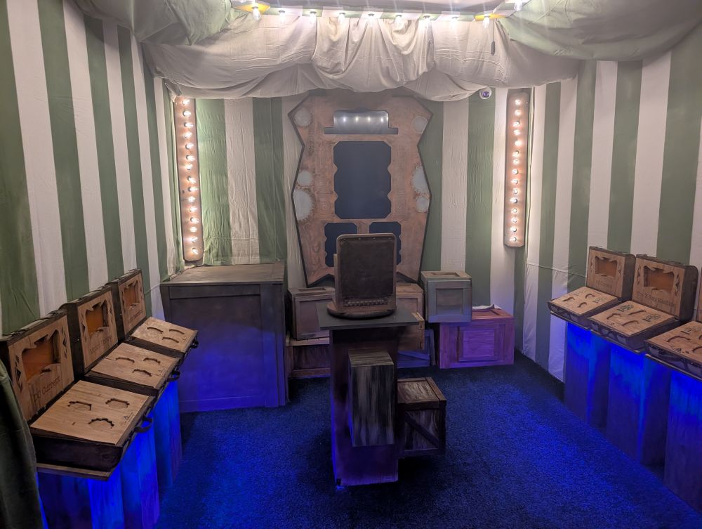 A room that appears to be a cross between a green-and-white-striped circus tent, a warehouse, and a video console of some sort. The far wall has a pile of boxes and the video screens. The side walls each have a series of three wooden briefcases with indentations for objects. The walls are draped in striped fabric, and several small light bulbs outline the corners and the center of the ceiling.