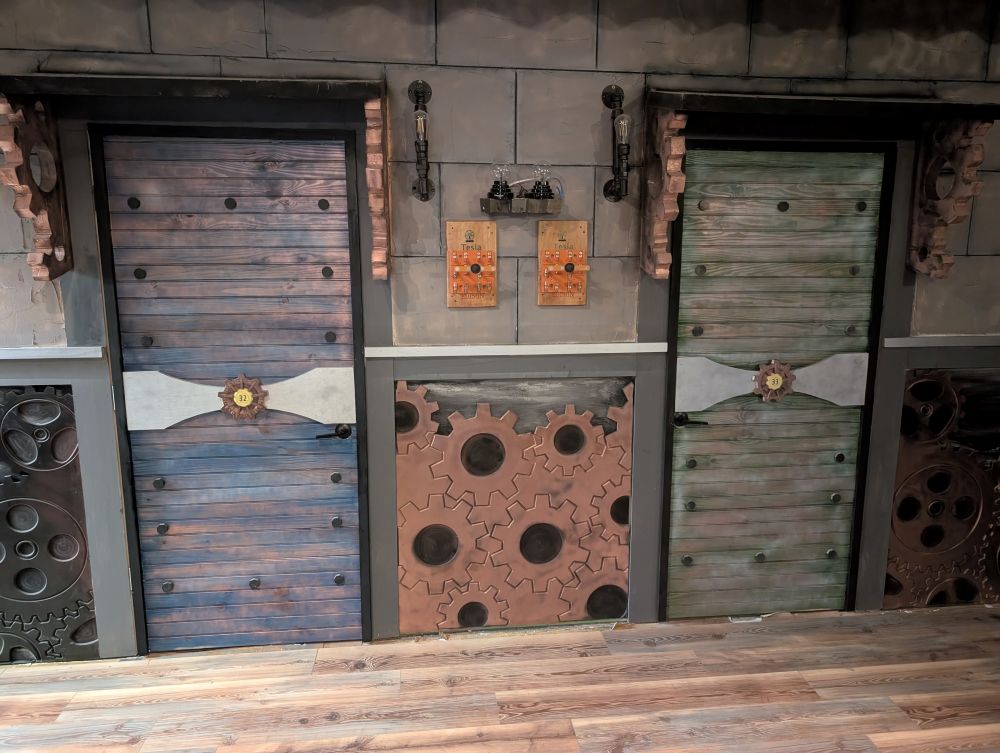A gear-filled wall with two identical warehouse doors. Between the doors are Edison-bulbed lights, two miniature plasma globes, and two large switches that can toggle between "Tesla" and "Edison." The scene has a steampunk vibe.
