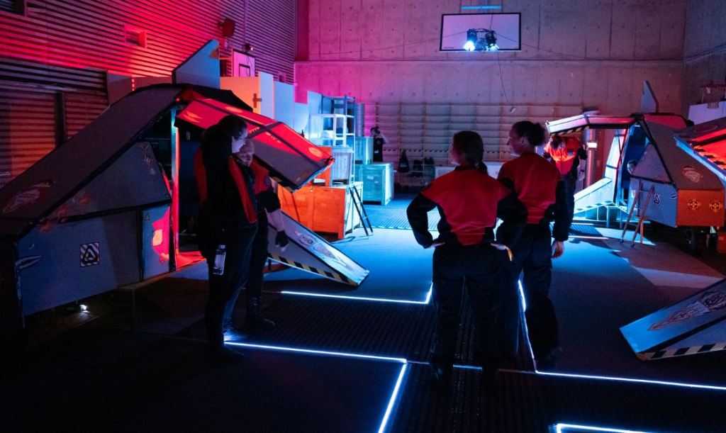 people in red crew uniforms standing in the set of a hanger bay, with spacecraft props