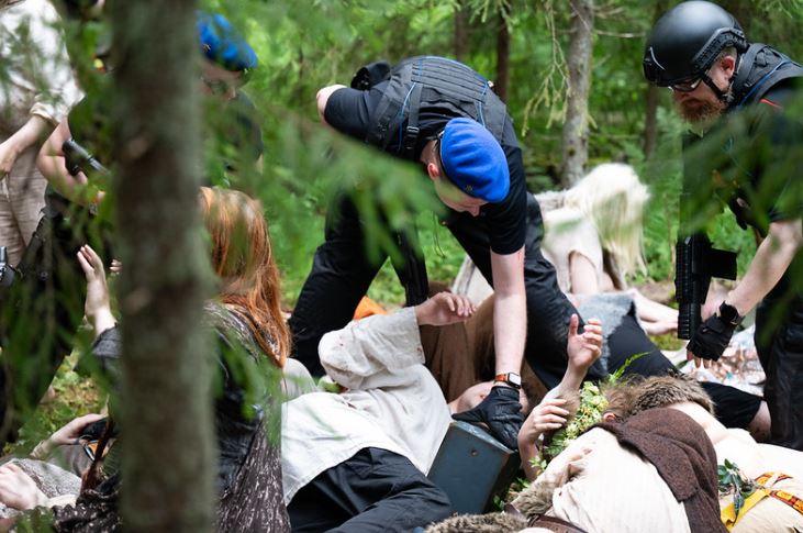 uniformed people standing over civilians in loose fitting clothing. the civilians are lying on the ground helpless. they are in the woods.