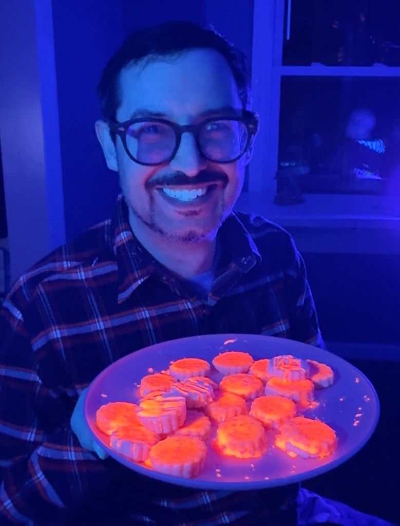 David holding a plate of overwhelmingly bright orange cookies glowing under black light.