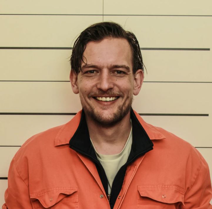 man in orange jumpsuit, medium brown hair parted on the side, smiling, standing against a wall with horizontal lines