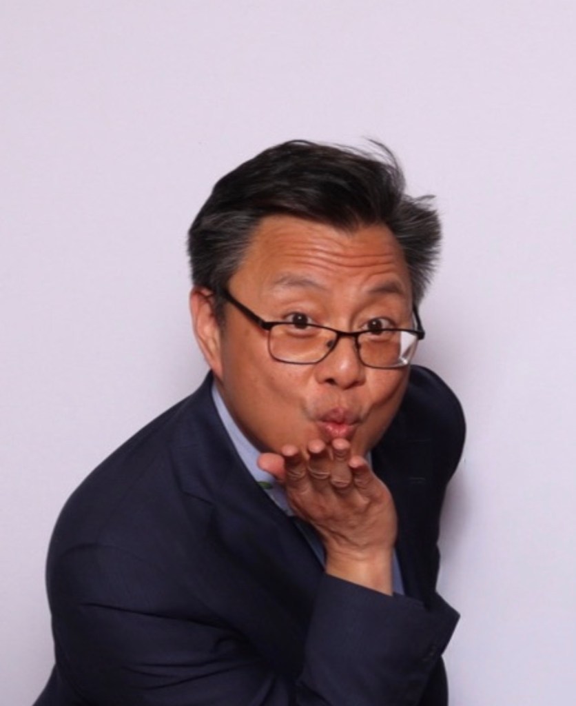 asian man wearing glasses and a suit, blowing a kiss at the camera