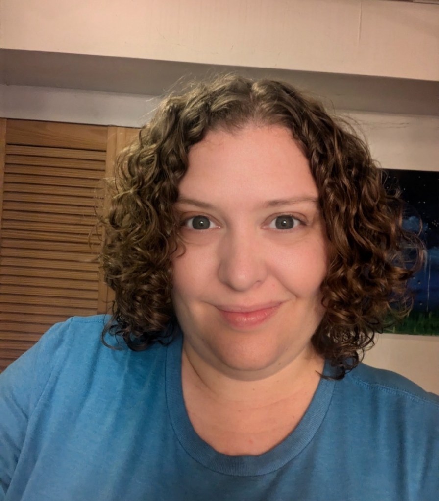 woman with grey eyes and shoulder length dark blonde curly hair smiling, in blue t-shirt