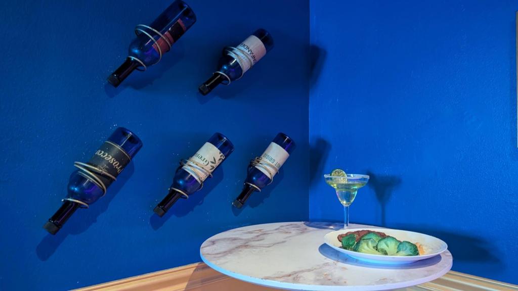 The corner of a room with a marble-topped table holding a drink and a plate of food. Wine bottles are in holders mounted to the wall.