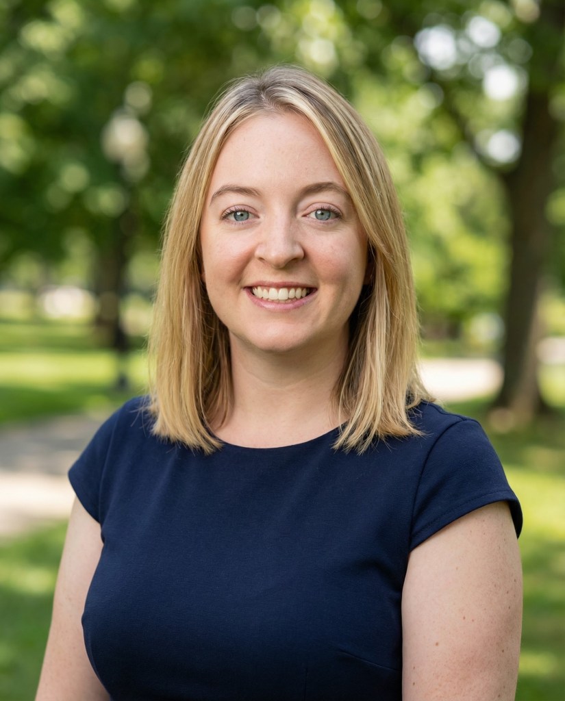 woman with shoulder length straight blonde hair and blue eyes, wearing a blue t-shirt, smiling.