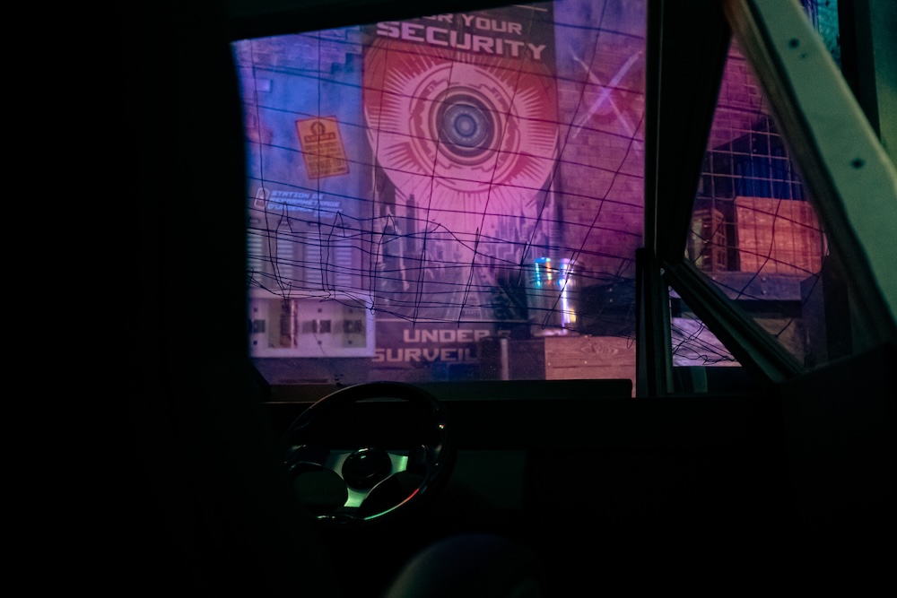 A view from inside of a vehicle of a cyber punk city with a large banner warning that everyone is under surveillance.