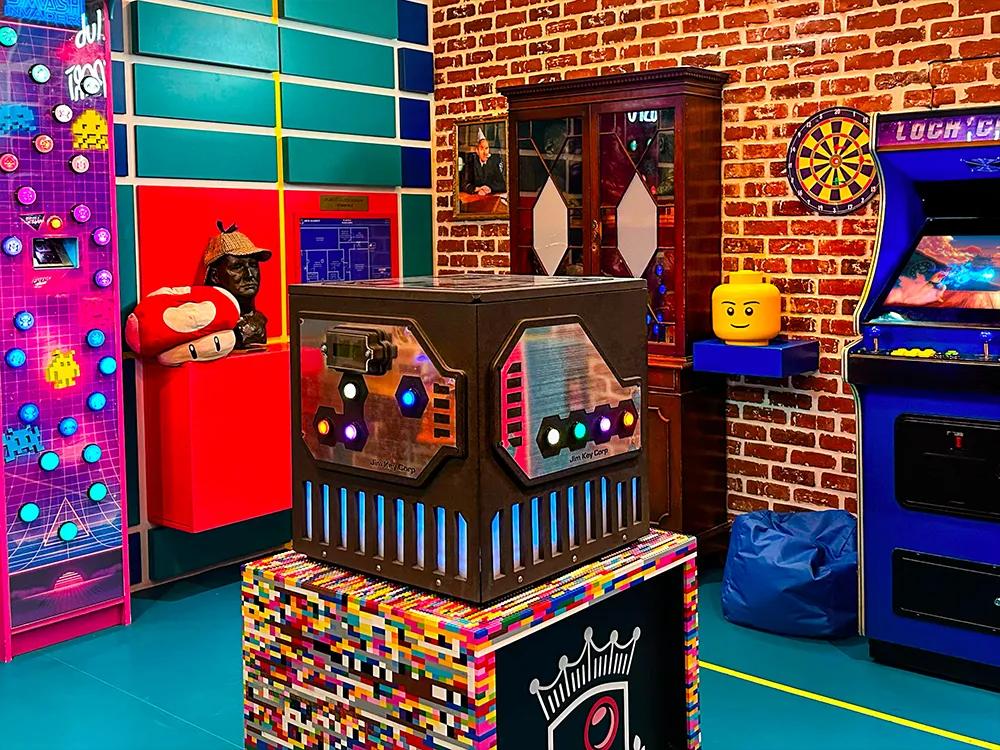 A colorful room with a large cube with glowing buttons on a LEGO pedestal, and various arcade games and shelves in the background