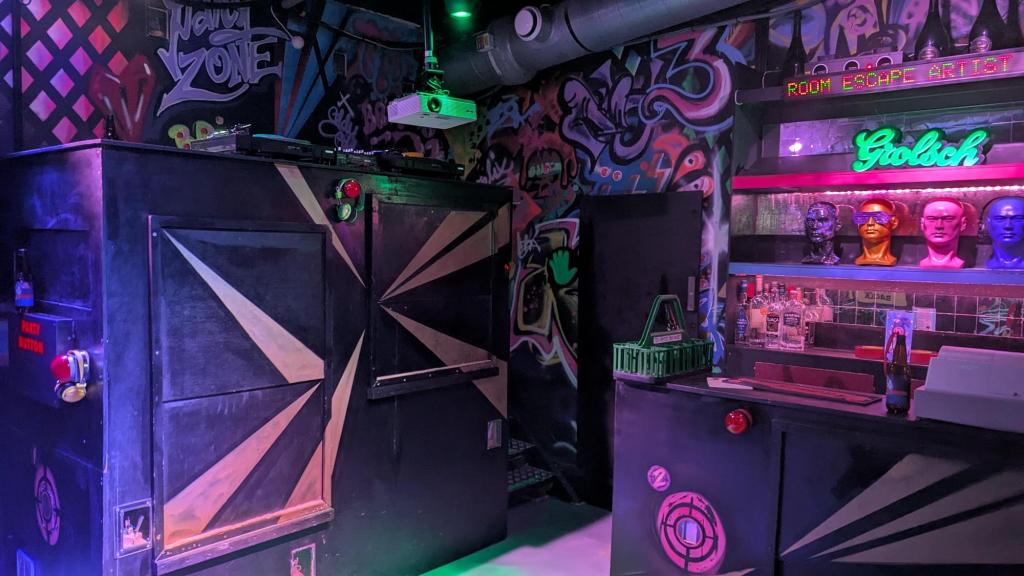 A grungy night club with colorful graffiti covering all the walls, and "Room Escape Artist" written on an LED display behind the bar