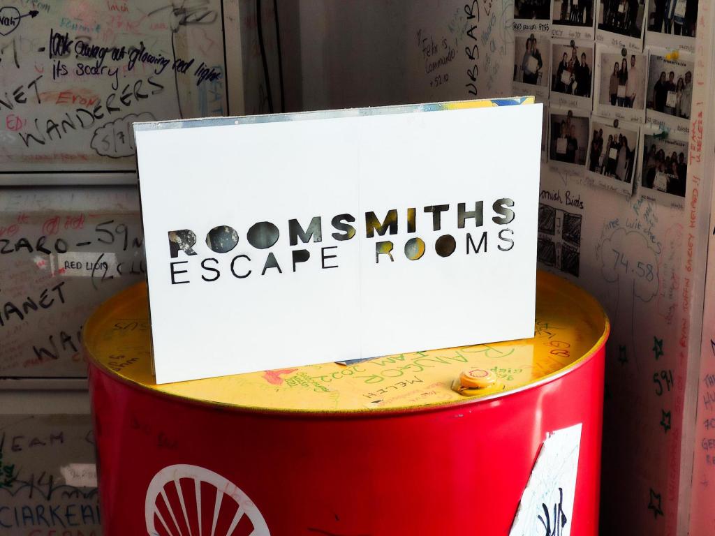 A sign that says "Roomsmiths Escape Rooms" placed on a red and yellow metal barrel