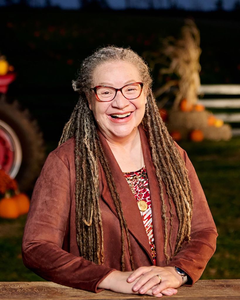 older woman with long grey and brown dreadlocks, cat eyed classes, smiling broadly, in a rust color blazer and red patterned top