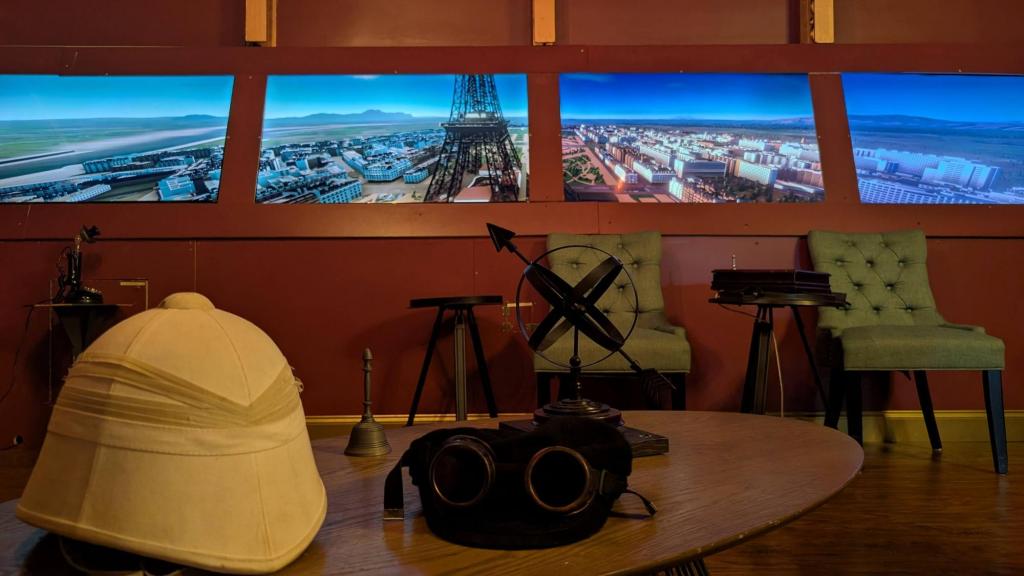 A pith helmet and some other old-time objects on a table, with a bank of windows in the background showing an aerial view of Paris.