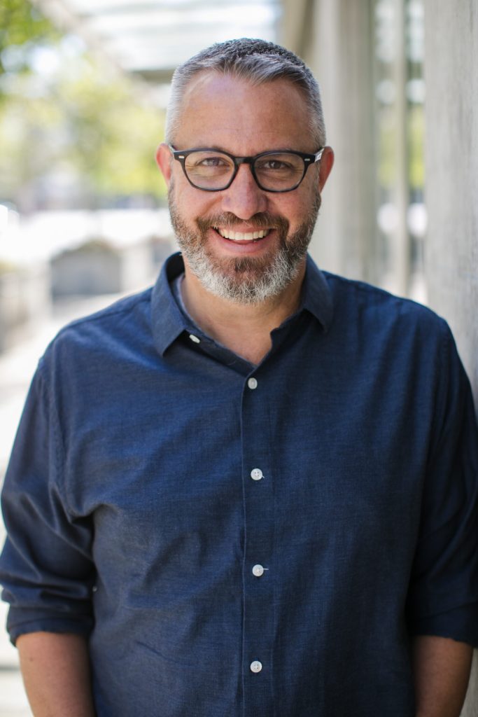 Man with salt and pepper hair and beard, smiling, wearing glasses and a dark blue button up shirt with sleeves rolled to elbows