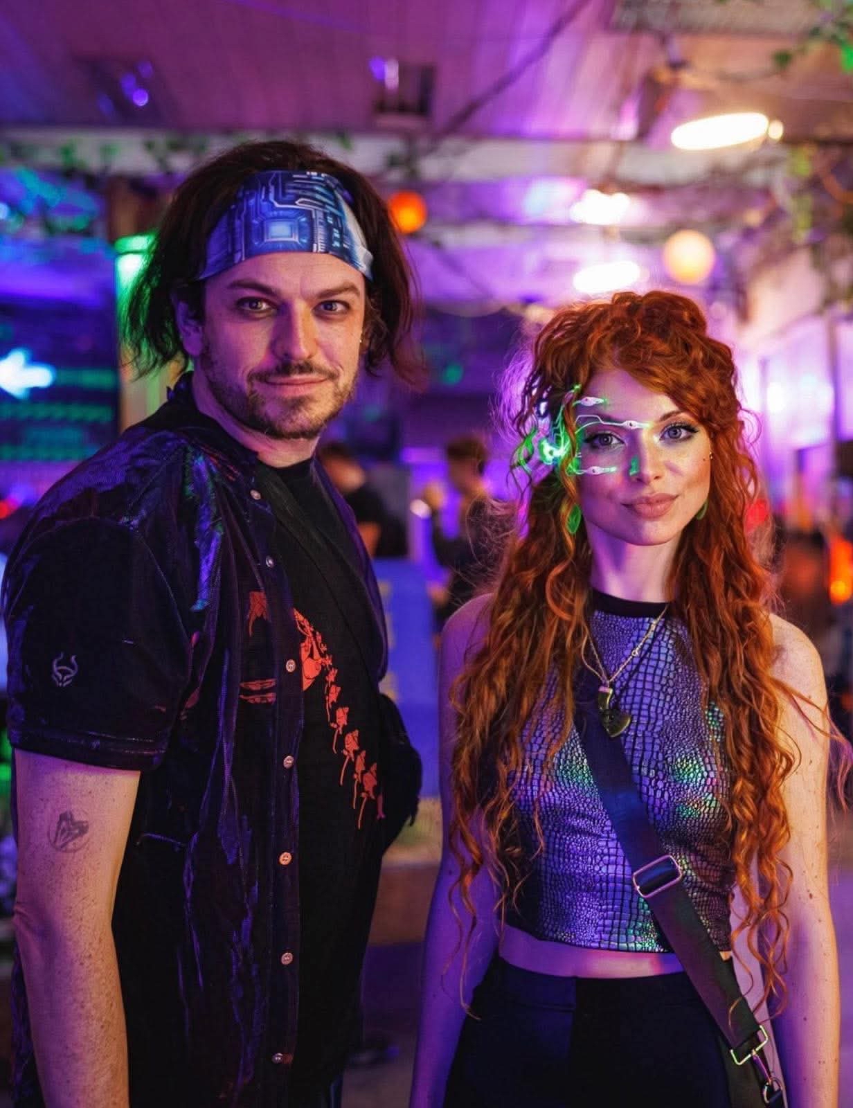 man with short beard and long dark brown hair, wearing a headband and shiny open shirt over a t-shirt. Woman with long curly red hair with a lit-up cyberpunk face accessory in a shiny halter top.