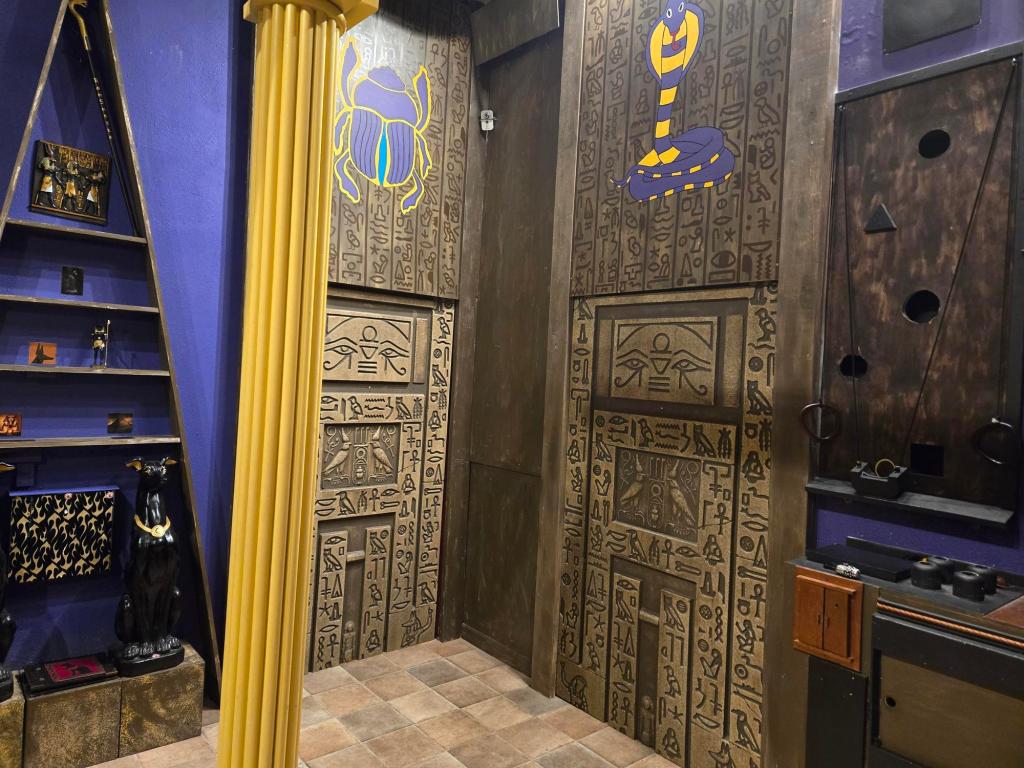 Two 15 foot tall panels covered in hieroglyphs that are flanked by a tall triangular set of shelves on the left with a dog statue at the bottom and pictures on the shelves and a desk to the right with a wall-mounted dexterity puzzle above it.