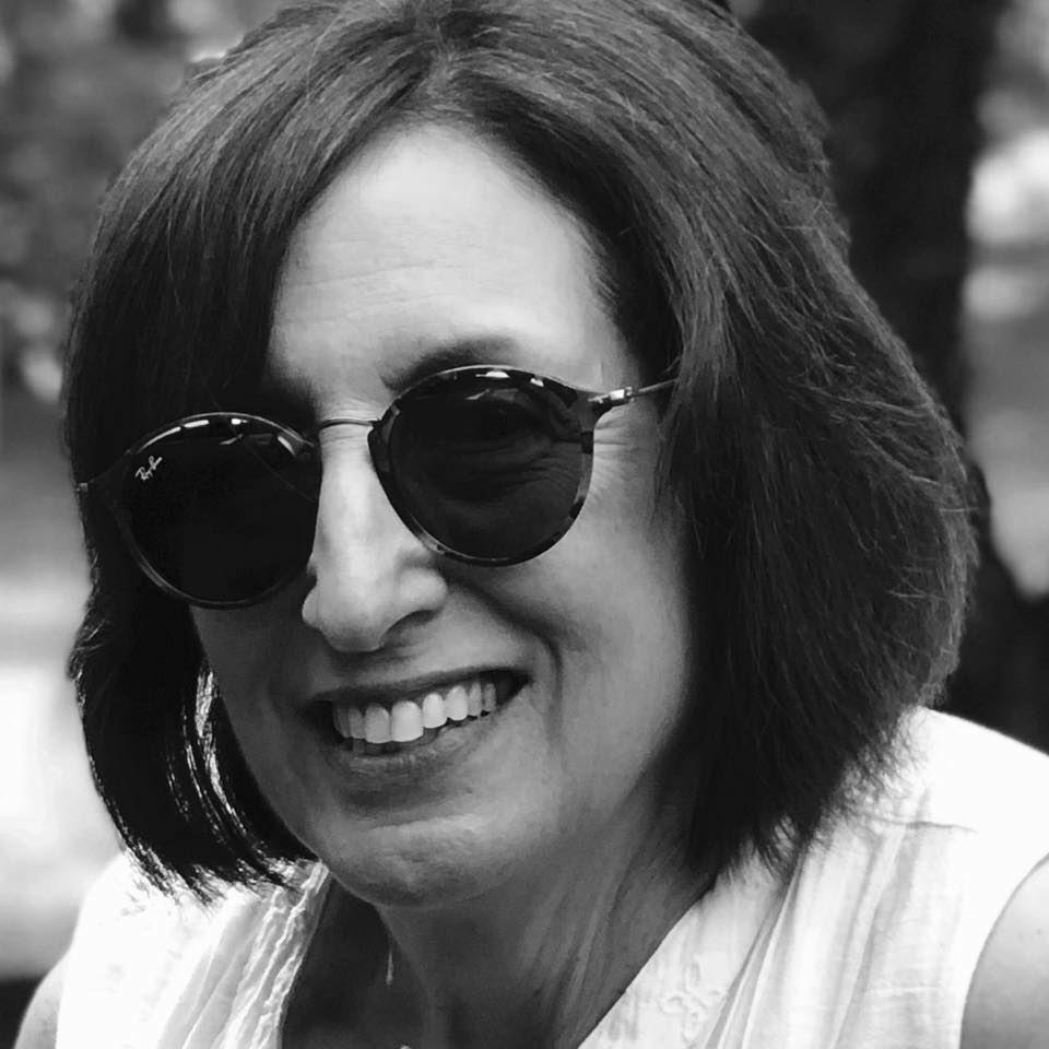 black and white image of woman with chin length dark hair, wearing sunglasses and a white top, smiling