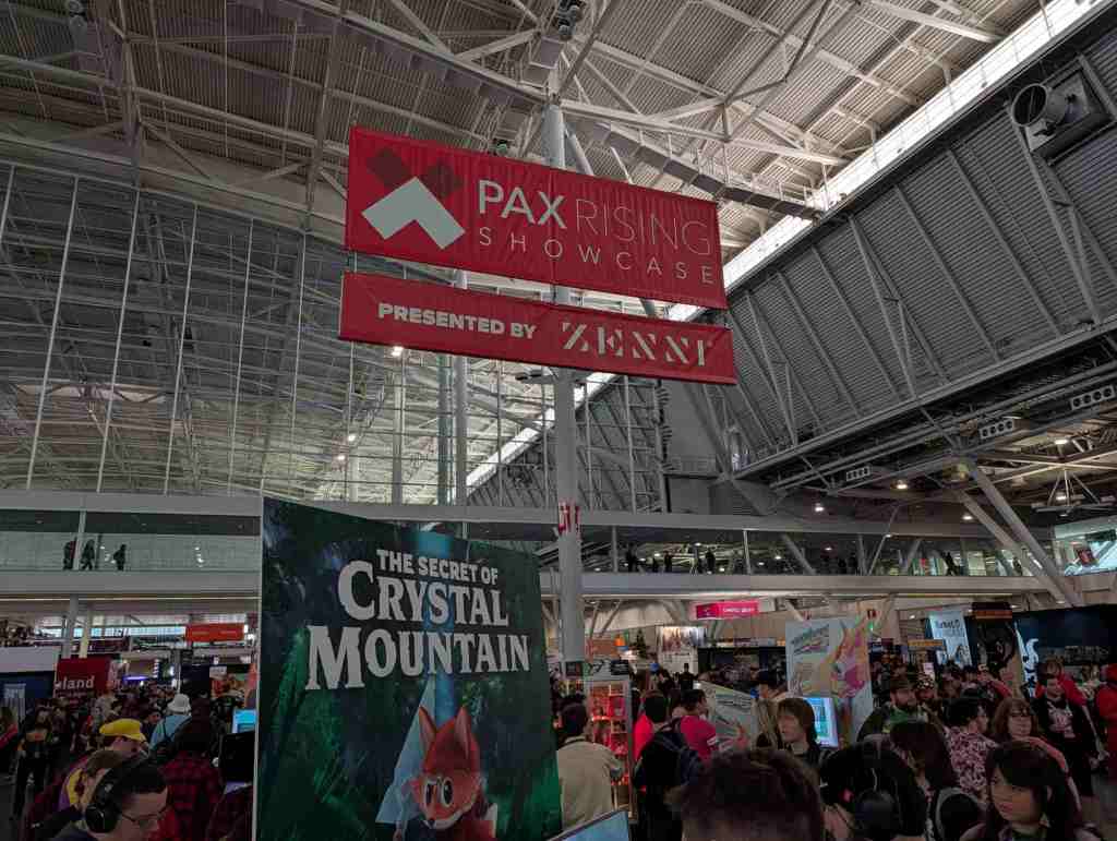 A sign hanging from a tall ceiling showing where the PAX Rising Show case is. Below, a poster for The Secret of Crystal Mountain.
