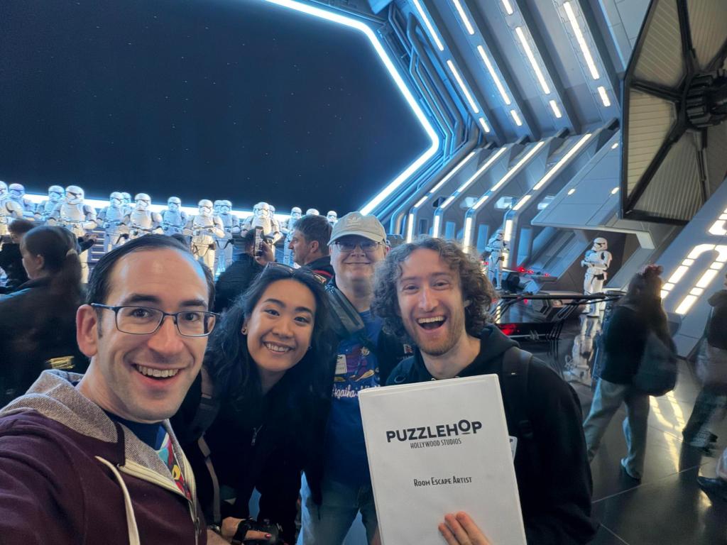 REA contributor Matthew Stein and team holding a "Puzzlehop" binder in front of the Stormtroopers on the Rise of the Resistance ride