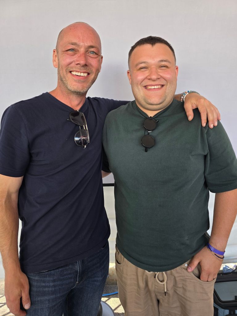 two men smiling at camera with their arms around each others shoulders. both are in plain dark color t-shirts with sunglasses hanging in the front.