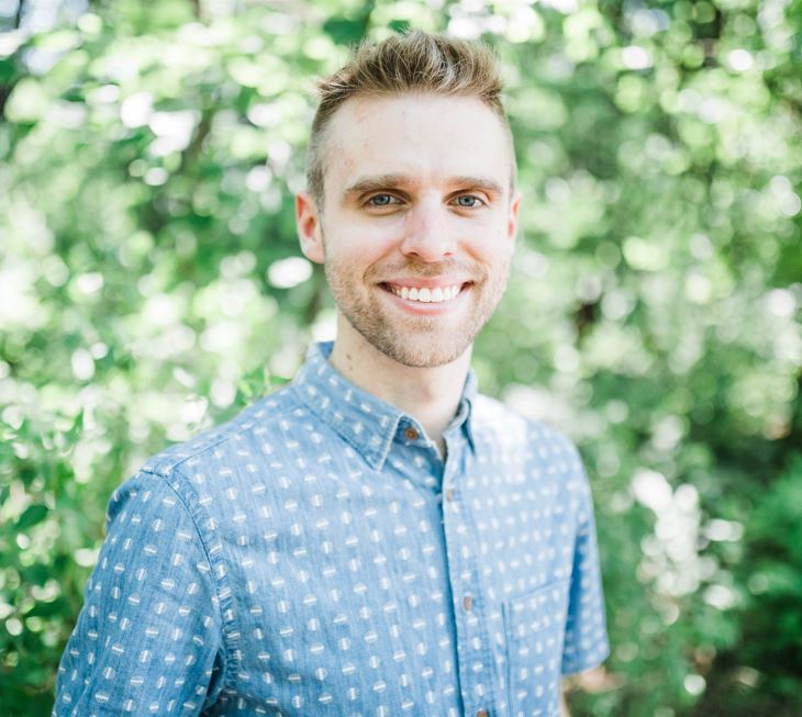 man with blonde hair, very short beard, blue eyes, smiling, in a button down patterned blue shirt.