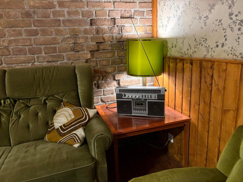 Corner of a room showing part of a green velvet couch, a green lamp on a wooden end table, The wall is brick, with wood paneling.