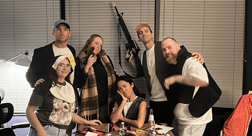 group of players wearing costume accessories such as nun's habit, flak vest, and holding toy guns. spread out on the table in front of them are booklets and papers from a game