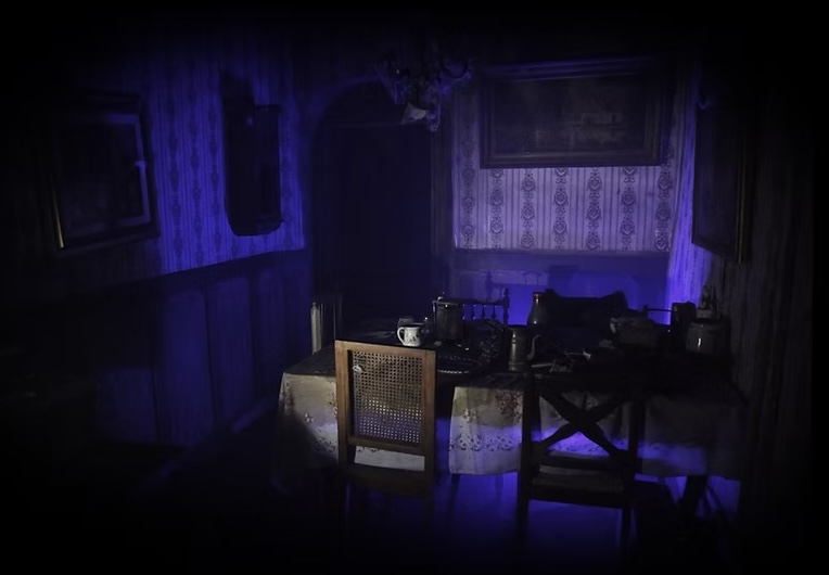 A table with a table cloth and place settings and 4 chairs around it sits int he center of a dark room with wallpapered walls with big, framed art hanging on each wall
