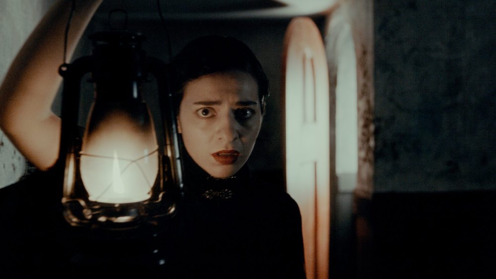 A woman dressed in black and holding a lantern stands in a hallway with a frightened look on her face.